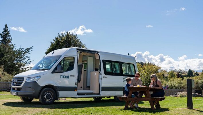 Een gezin zit aan een picknicktafel voor de Maui Ultima Plus camper die geparkeerd staat op het gras