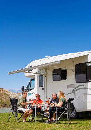 De zijkant en achterkant van de Maui River camper waar een familie onder de luifel zit aan tafel op de campingstoelen en een kind vanuit de zithoek achterin de camper naar buiten kijkt