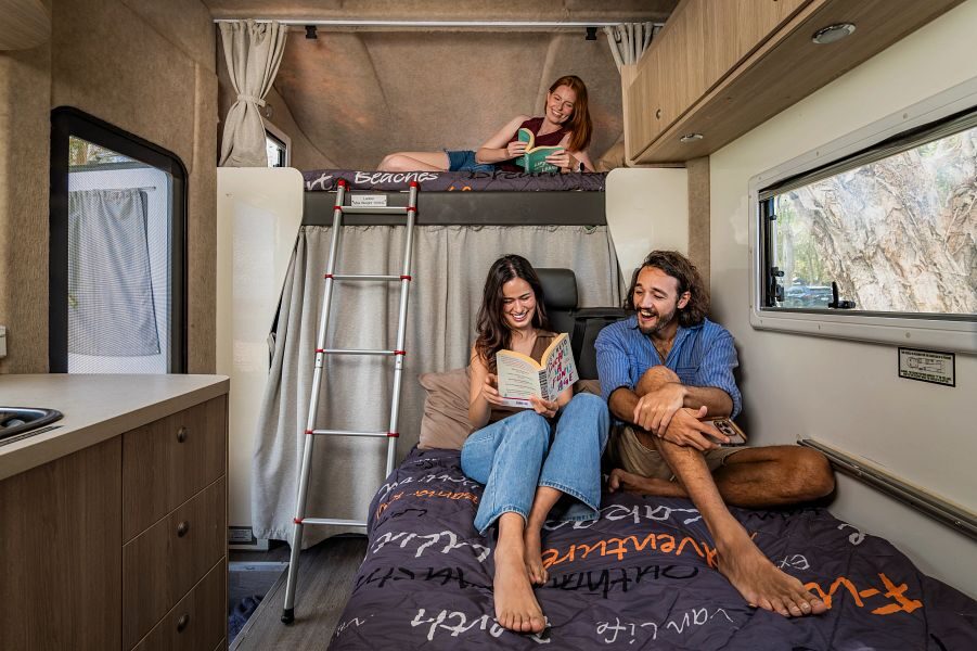 Een koppel zit op het bed in het midden van de Mighty Big Six camper een boek te lezen en achter hun ligt een dame op het tweepersoonsbed in de alkoof