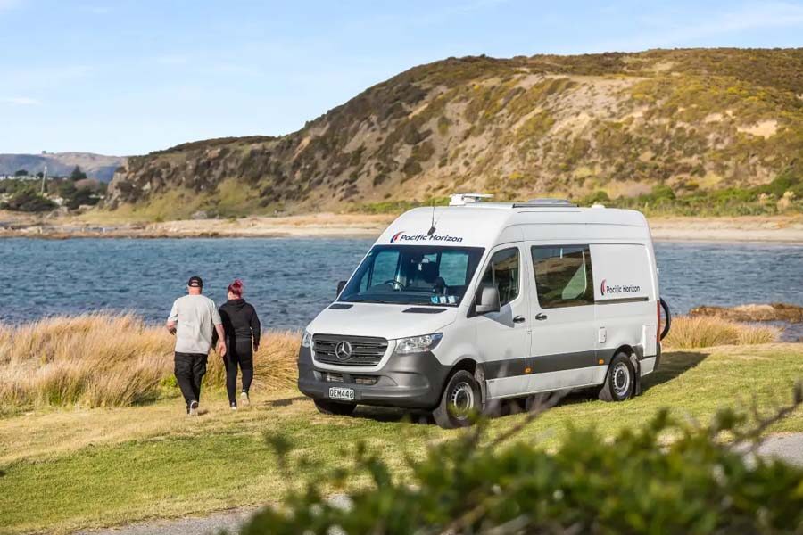 Een koppel loopt vanuit de Pacific Horizon 2 plus 1 camper naar een meer toe in Nieuw-Zeeland