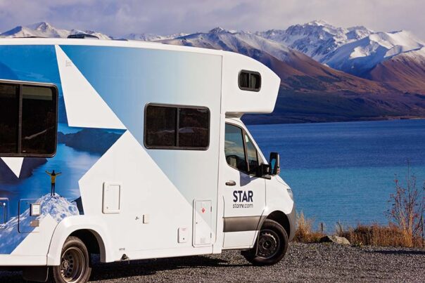 De zijkant van de Star RV Polaris 6 camper die geparkeerd staat naast een meer met uitzicht op besneeuwde bergtoppen