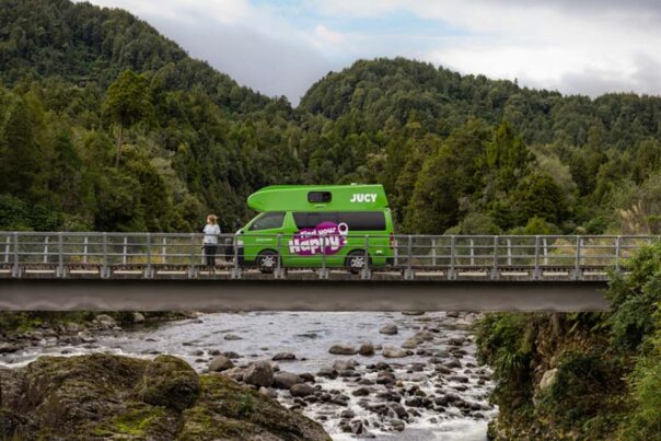 Een vrouw staat naast de op een brug geparkeerde Jucy Chaser camper in Nieuw-Zeeland